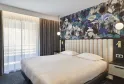 Chambre d'hôtel moderne avec lit queen-size, tête de lit capitonnée crème, papier peint floral bleu et blanc, grande baie vitrée avec rideaux, luminaires suspendus et mobilier contemporain.