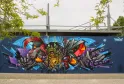 Fresque de street art colorée sur le M.U.R de Bordeaux représentant des visages stylisés et des créatures fantastiques avec des teintes bleues, rouges, or et violettes, sous un pont moderne.