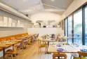 Intérieur moderne d'un restaurant avec tables en bois clair, chaises jaunes et marron, grandes baies vitrées donnant sur une terrasse, plafond blanc avec poutres apparentes et étagères décoratives.