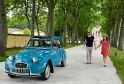 Une Citroën 2CV bleu turquoise garée sous une allée d'arbres à Bordeaux, avec un couple se promenant à côté et des bâtiments classiques en arrière-plan.
