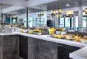 Buffet de petit-déjeuner moderne avec comptoir noir et blanc présentant fruits frais, viennoiseries et plats variés, dans une salle vitrée avec éclairage contemporain à l'Hôtel Au Comté d'Ornon à Bordeaux.
