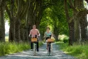 Deux cyclistes qui pédalent sur une allée de gravier bordée de grands platanes, avec des paniers en bois sur leurs vélos, dans les vignobles de Bordeaux.