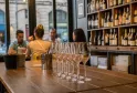 Verres à vin alignés sur le comptoir d'une cave à vin bordelaise, avec des étagères de bouteilles en arrière-plan et des clients dégustant du vin.
