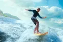 Surfeur en combinaison noire chevauchant une vague avec équilibre, sous un ciel bleu dégagé avec des montagnes verdoyantes en arrière-plan
