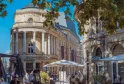 Place de la Comédie à Bordeaux avec le Grand Théâtre aux colonnes majestueuses, les bandeaux Cinémas, terrasses avec parasols, et feuillage d'automne sous un ciel bleu.