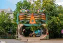 Installation artistique colorée représentant un visage stylisé avec des yeux turquoise et des motifs géométriques orange et violets, formant une arche d'entrée à un tunnel dans un espace vert alternatif de Bordeaux.