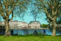Vue de la Garonne à Bordeaux avec des bâtiments historiques en arrière-plan, des arbres centenaires au premier plan, et des personnes pratiquant des loisirs au bord de l'eau.