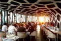 Restaurant gastronomique à Bordeaux avec plafond géométrique blanc et rouge, tables élégantes avec couverts, clients en tenue formelle dînant, grandes baies vitrées avec vue sur la ville au coucher de soleil.