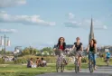 Trois femmes à vélo sur une piste cyclable dans un parc de Bordeaux, avec la basilique Saint-Michel visible en arrière-plan et des promeneurs sur l'herbe.