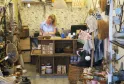 Une femme travaille dans une petite boutique artisanale remplie de paniers tissés, de décoration et de produits naturels, avec des murs décorés de papier peint motifs fleuris.
