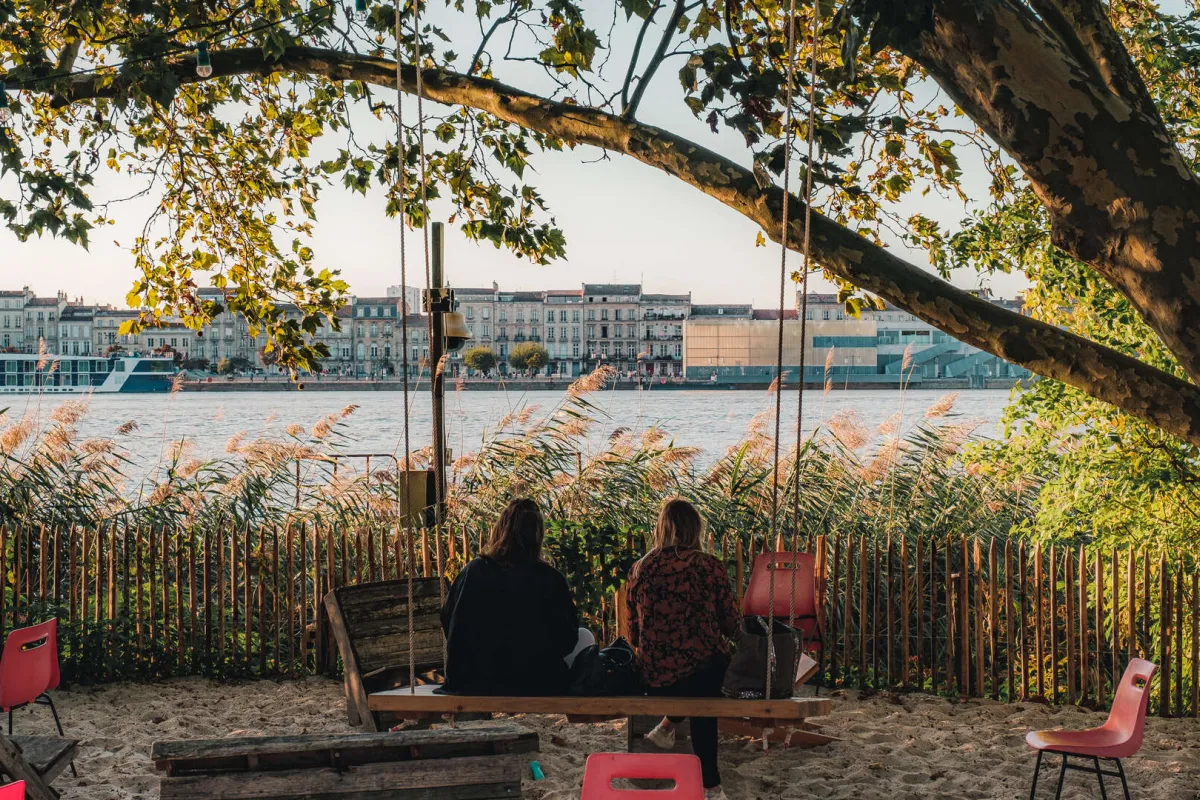 Deux personnes assises sur un banc sous un grand arbre au bord de la Garonne à Bordeaux, avec vue sur les façades historiques et les bâtiments modernes de la ville de l'autre côté du fleuve.