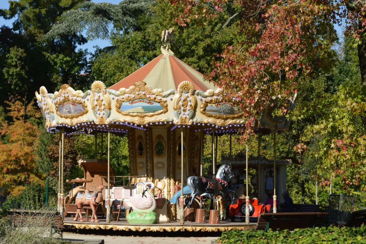 Un carrousel vintage aux couleurs blanc et or avec un toit rouge et orange, entouré d'arbres aux feuillages d'automne rouges et dorés dans un parc de Bordeaux.