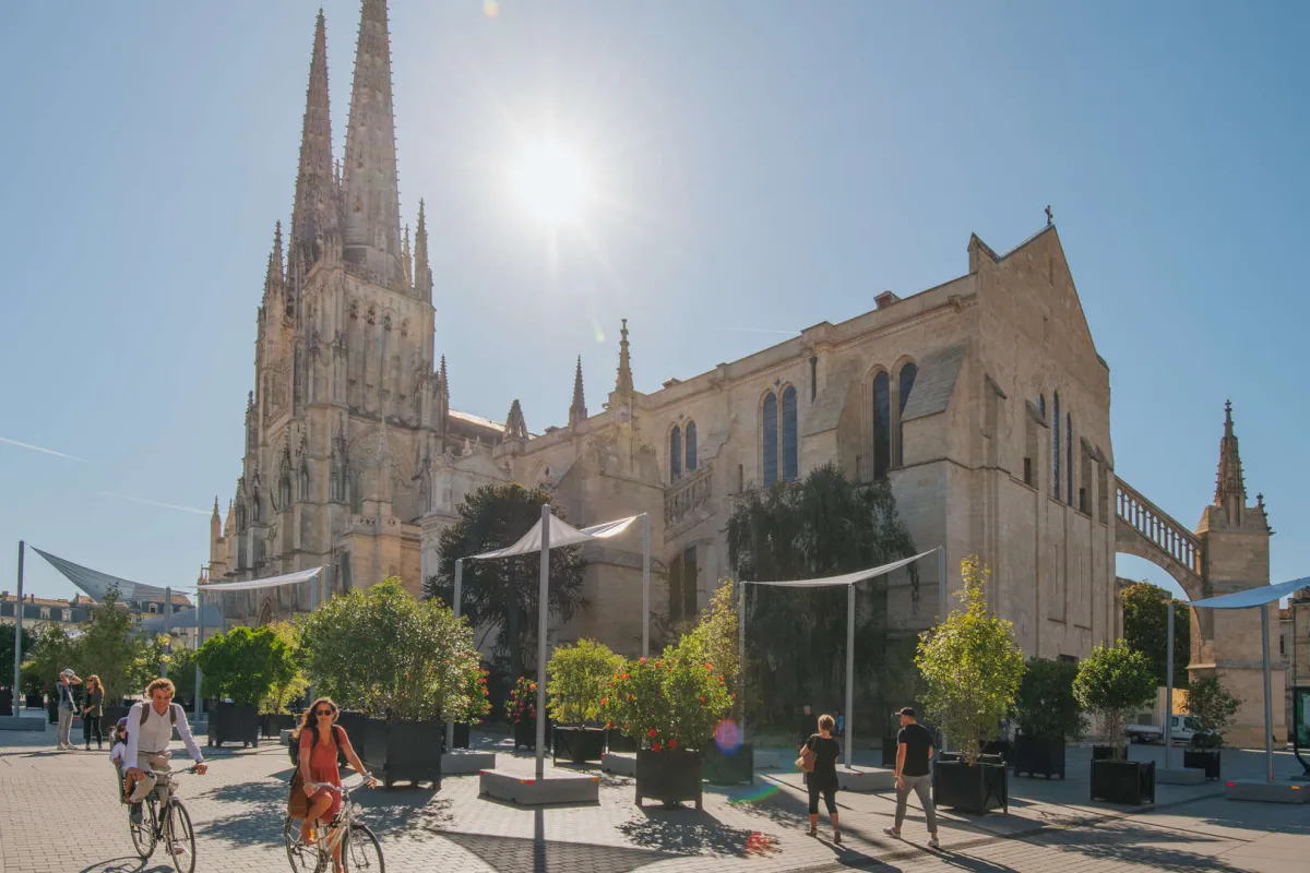 Place de la Cathédrale à Bordeaux avec la Basilique Saint-Michel en arrière-plan, des cyclistes et des piétons circulant dans l'espace public aménagé avec des voiles d'ombrage et de la végétation.