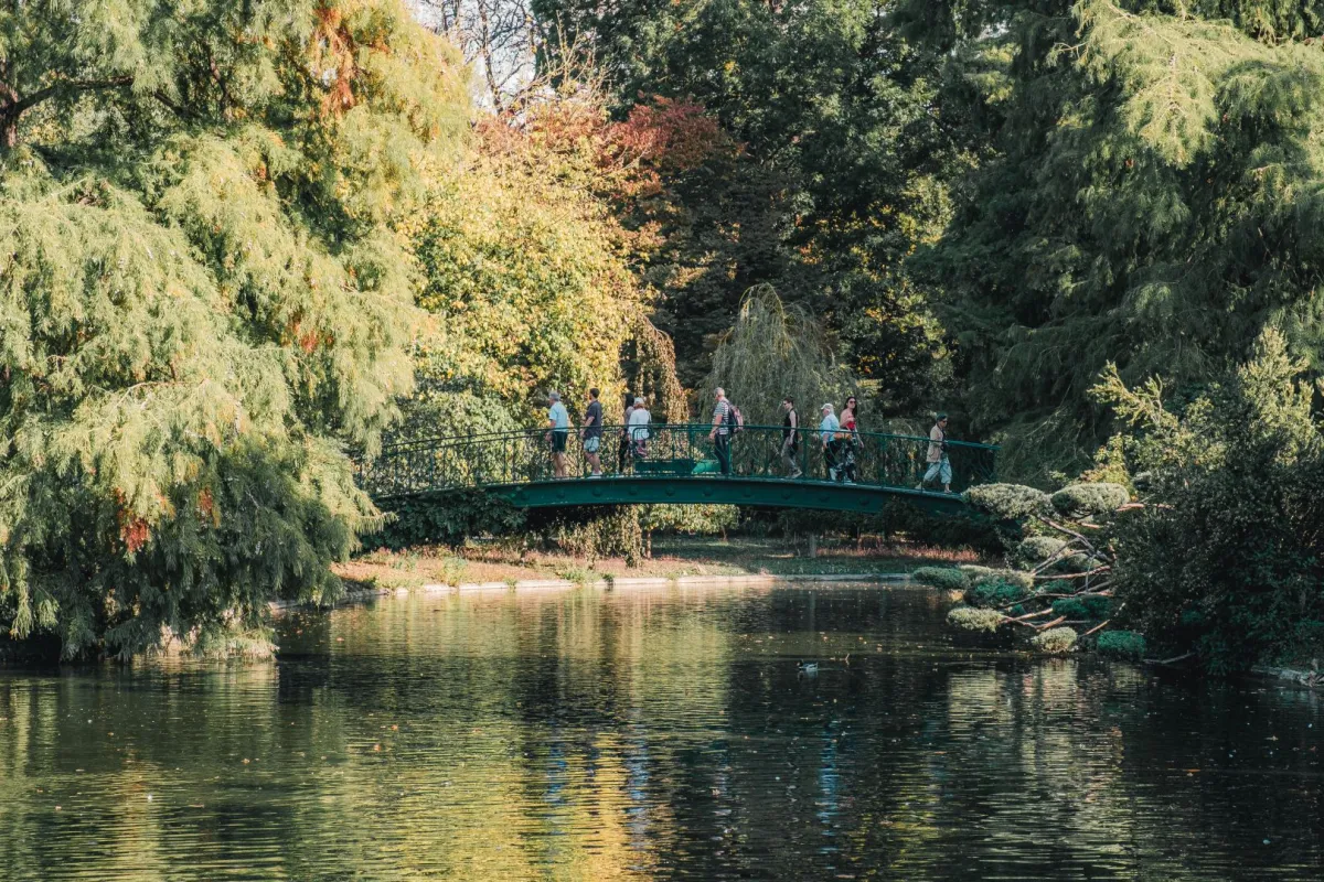 Un groupe de visiteurs traverse un pont vert arqué au-dessus d'une rivière calme, entouré de saules pleureurs et d'arbres aux feuillages d'automne dans un parc de Bordeaux.