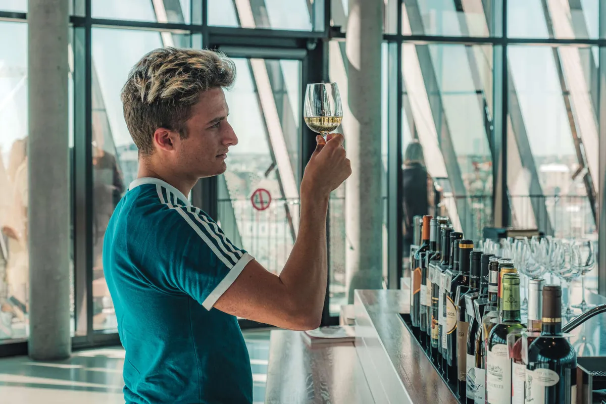 Jeune homme en chemise bleue dégustant un verre de vin blanc dans un bar à vin de Bordeaux, avec vue panoramique sur la ville depuis une baie vitrée.