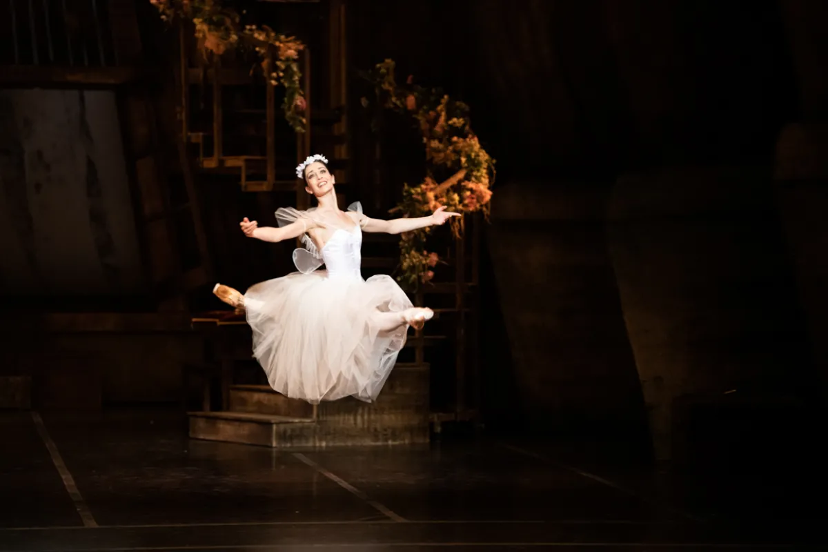 Ballerine en tutu blanc effectuant un saut en arabesque sur scène, les bras écartés, avec un décor architectural doré en arrière-plan.