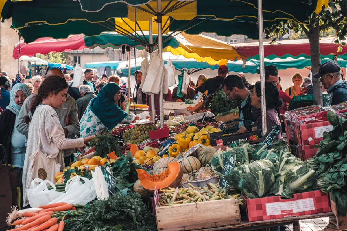 Marché en plein air à Bordeaux avec vendeurs et clients sous des parasols colorés verts, jaunes et rouges, étalant des fruits et légumes frais dont des courges, poivrons, carottes et légumes verts.