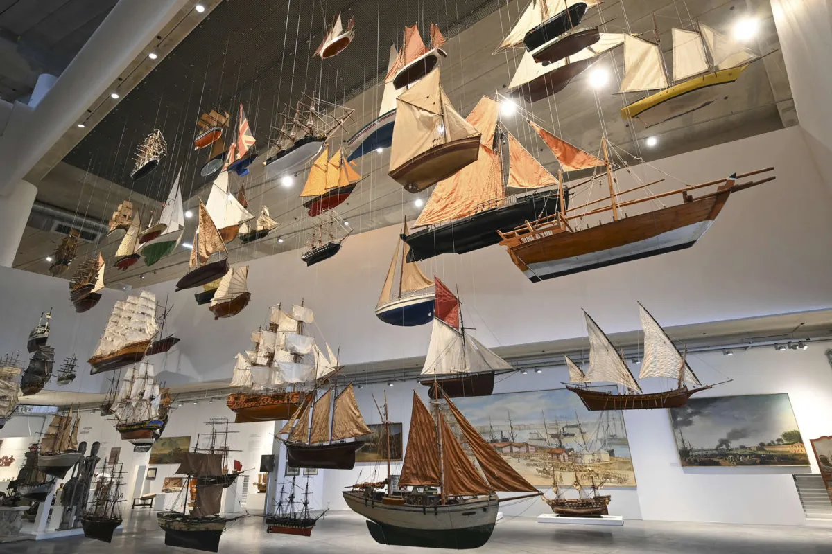 Collection de maquettes de navires historiques suspendus au plafond d'un musée, représentant différents types de bateaux à voiles des XVIIe et XVIIIe siècles, avec des tableaux maritimes exposés sur les murs blancs.