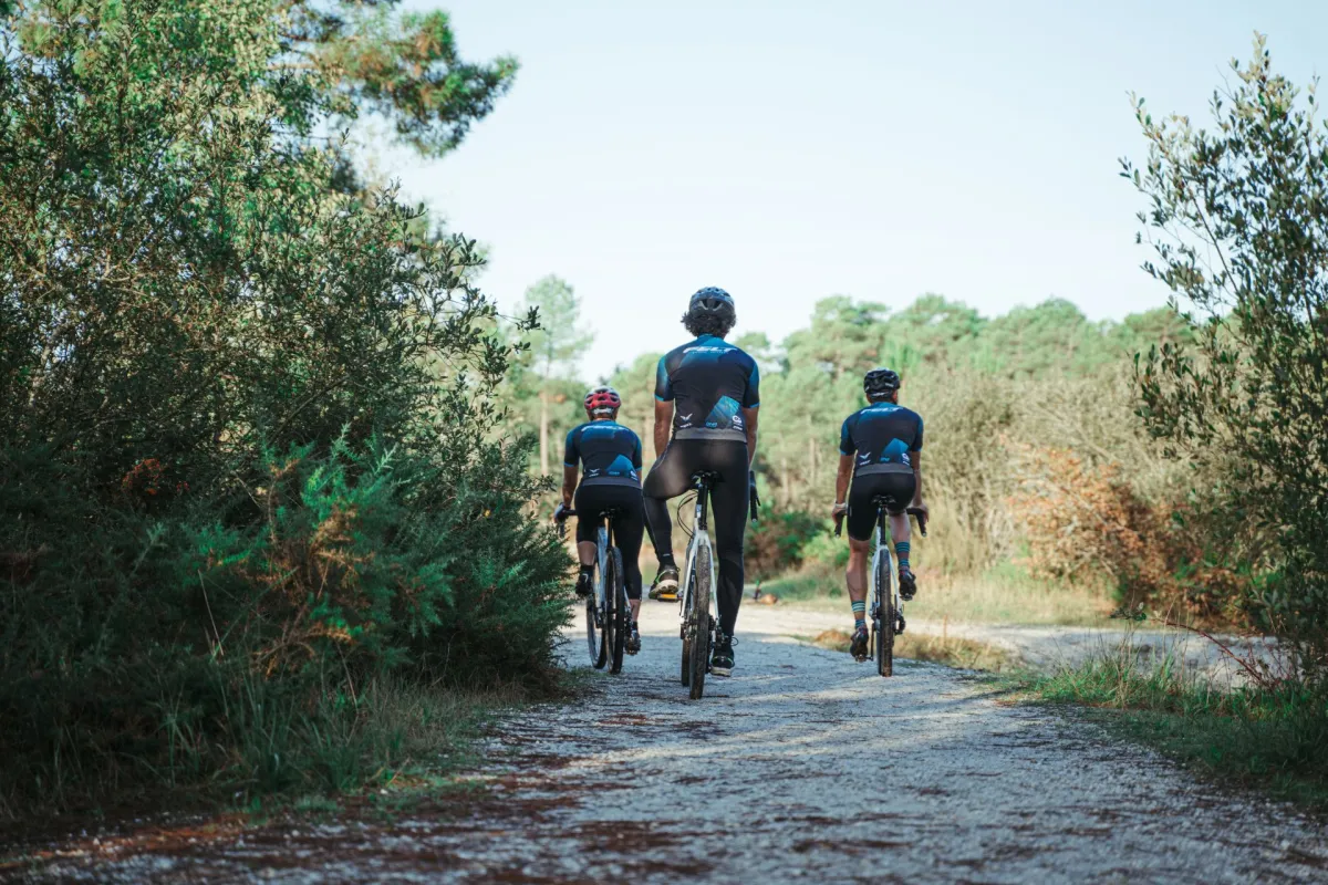 Trois cyclistes en tenue de sport bleue et noire circulant sur un chemin de gravel bordé de végétation méditerranéenne