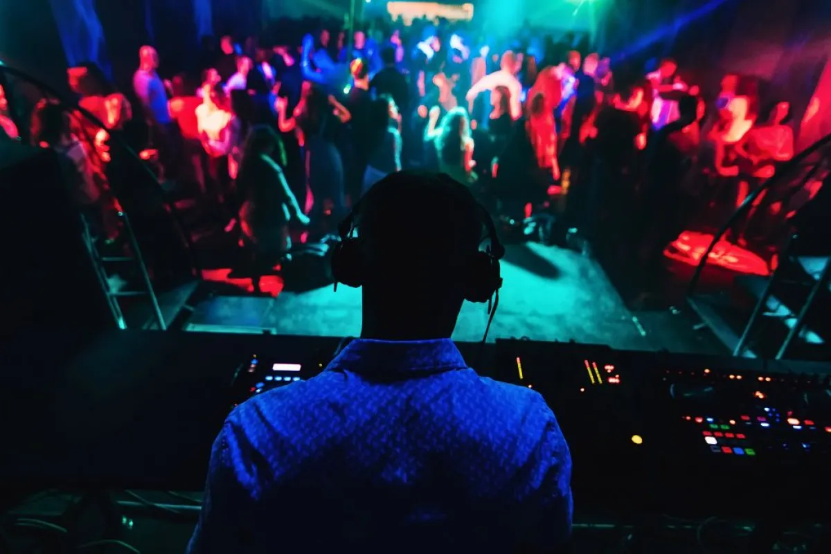 DJ en silhouette portant un casque derrière sa console de mixage, face à une foule dansante éclairée par des lumières rouges et bleues dans une boîte de nuit.