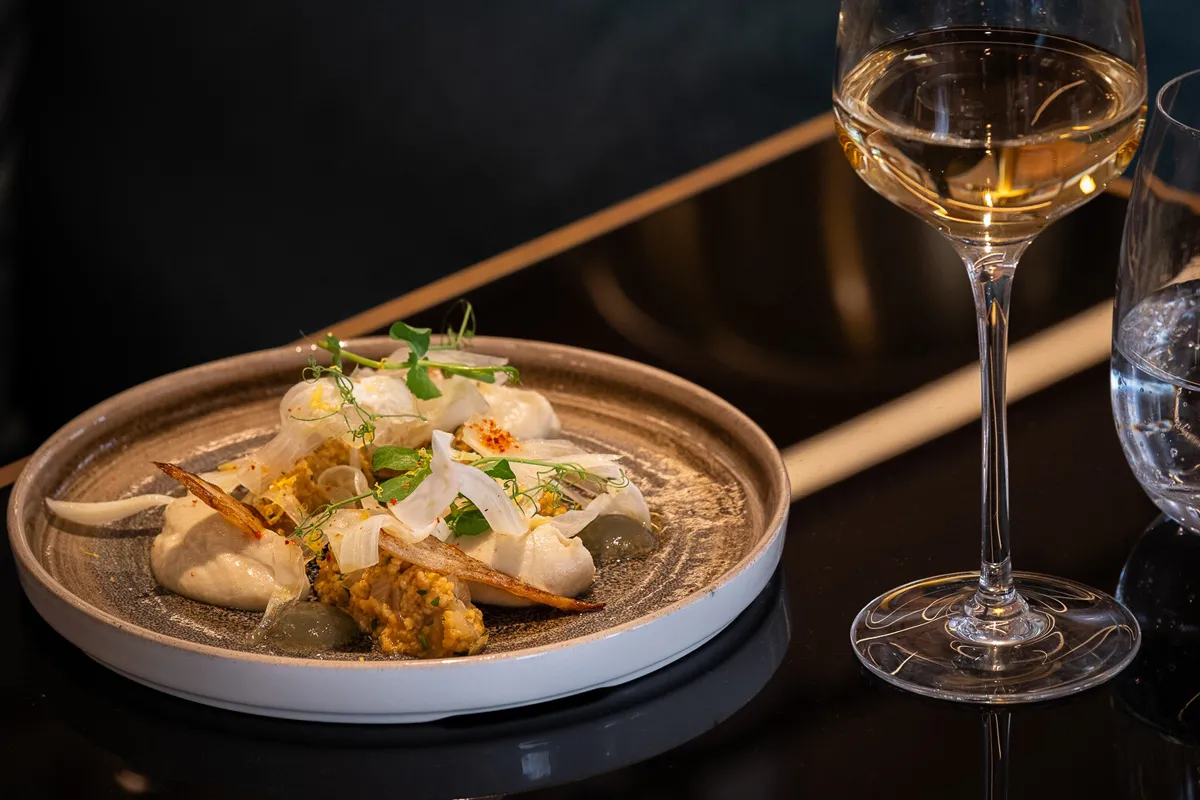 Assiette gastronomique présentant des noix de Saint-Jacques, légumes rôtis et microherbes, accompagnée d'un verre de vin blanc sec sur une table noire